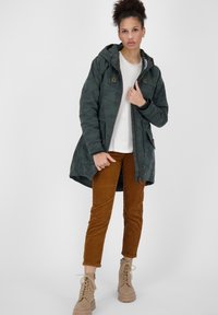 Parka camouflage verde con cappuccio, vita regolabile con cordoncino e due tasche frontali, abbinata a pantaloni in velluto corduroy marroni e stivali beige con lacci.