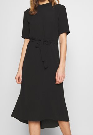 Woman wearing a knee-length black dress with short sleeves and a tied belt at the waist, standing against a plain light gray background.