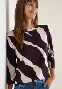 Maglietta a maniche lunghe di colore viola scuro con strisce astratte beige. Realizzata in tessuto morbido, presenta un collo a giro e una vestibilità morbida, abbinata a dei jeans.