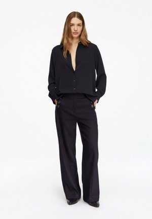 Kobieta stojąca z rękami w kieszeniach, ubrana w luźną czarną koszulę na guziki i szerokie czarne spodnie na białym tle.