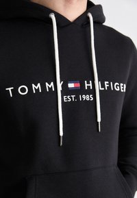 Zwarte hoodie gemaakt van katoen, met witte trekkoorden met metalen punten en een geborduurd logo met rode en blauwe accenten.