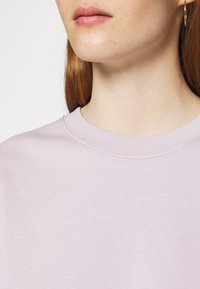 El sudadera de color lavanda claro tiene un cuello redondo. La tela parece suave con una textura lisa y un mínimo detalle de costura.
