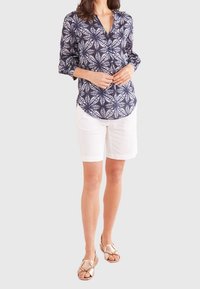 Blusa blu navy con bottoni e motivo floreale bianco, maniche lunghe arrotolate, abbinata a pantaloncini bianchi e sandali dorati.