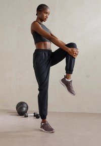 Černý sportovní top a volné černé tepláky s elastickými manžetami. Model nosí šedé sportovní boty a provádí cvik na zvedání kolen. Hmotnosti jsou poblíž.