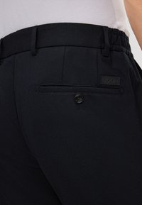 Close-up van zwarte broek met elastische tailleband, riemlussen en een achterzak met knoop en een klein rechthoekig merklabel.
