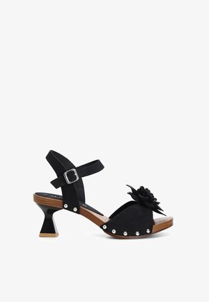 Black suede sandal with a floral accent, open toe, ankle strap, and silver studs. The heel is block-shaped with a wooden and glossy finish.