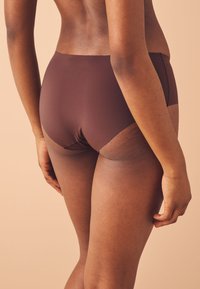 Primer plano de una persona que lleva ropa interior marrón sin costuras, mostrando la parte baja de la espalda, las caderas y la parte superior de las piernas sobre un fondo beige.