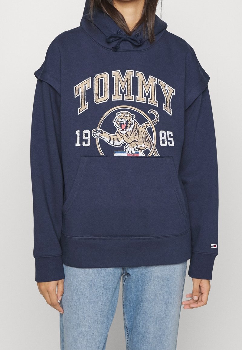 Tmavě modrá mikina s klokaní kapsou, grafikou lva, textem "TOMMY" a stahovací kapucí. Obsahuje žebrované manžety a lem, vyrobena z bavlněné směsi.