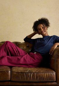 Tmavomodrá košeľa s dlhým rukávom skombinovaná s nohavicami s pruhmi v ružovej a burgundskej farbe, posedenie na hnedom koženom gauči, uvoľnená póza. Textúrované pozadie.