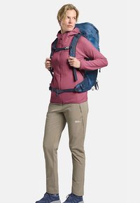 Blauer Wanderrucksack mit verstellbaren Riemen, getragen über einer pinken Jacke mit Reißverschluss und beigen Hosen. Robuste Schuhe, praktisches Design.