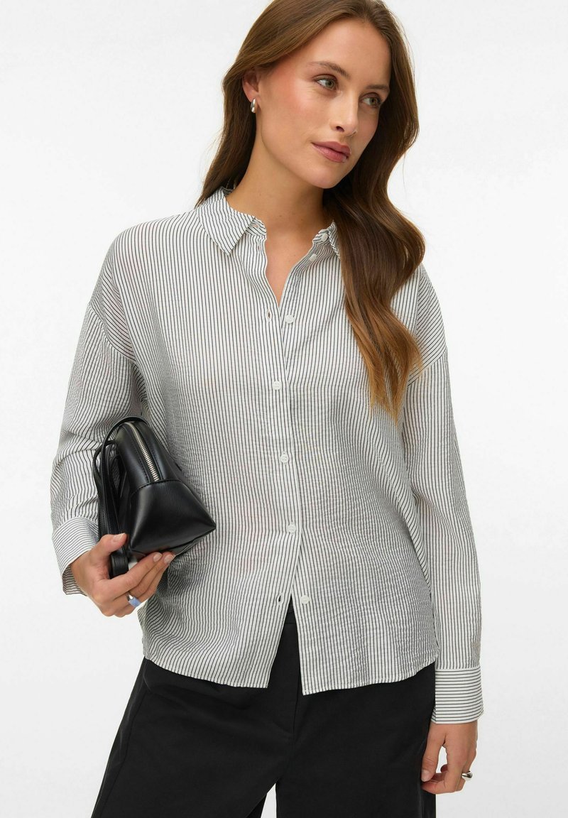 Femme portant une chemise blanche à rayures fines et un pantalon noir, tenant un sac à main en cuir noir avec une fermeture éclair.