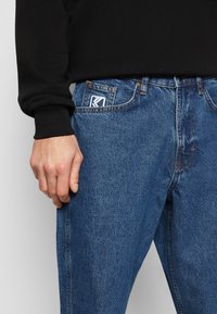 Blå denimjeans med lige ben, der har en frontlomme udsmykket med et hvidt logo-patch og kontraststing.