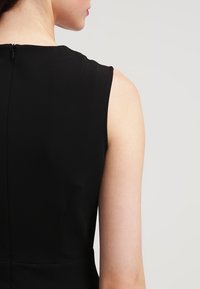 Schwarzes Kleid mit tailliertem ärmellosem Design, strukturierter Stoff und einem verdeckten Reißverschluss auf der Rückseite, der eine klare und moderne Silhouette präsentiert.