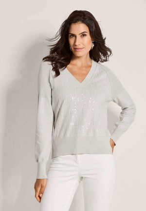 Frau mit welligem dunklem Haar, die silberne Ohrringe, einen hellgrauen V-Ausschnitt-Pullover mit dezenten Pailletten und weiße Hosen trägt, steht mit einer Hand in der Hüfte.