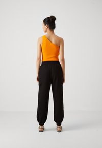 Naine seisab seljaga vaatajale, kandes oranži ühe õlaga varrukateta särk, mustad elastse vöökohaga joggerpüksid ja sandaalid ning tema juuksed on tehtud pundis.