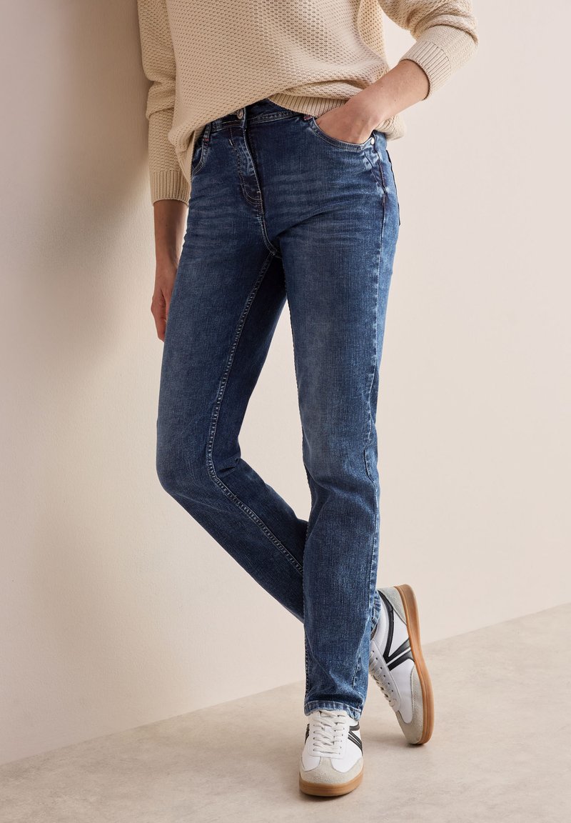 Blå denimjeans med slim fit, med subtil blekning, klassisk femfickdesign, kombinerat med vita sneakers och en krämfärgad tröja.