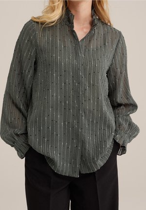 Woman wearing a sheer dark green blouse with vertical stripes and small dots, paired with black pants, standing against a beige background.