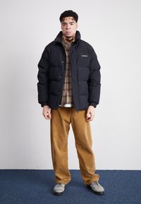 Schwarze Steppjacke mit Reißverschluss, gerippten Bündchen und dezentem Logo. Darunter ein gestreiftes Hemd in Schichten, kombiniert mit braunen Cordhosen und grauen Schuhen.