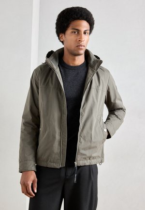 Veste zippée vert olive avec capuche, présentant une texture lisse, des poches avant, et un pull gris en dessous. Un pantalon noir est visible.
