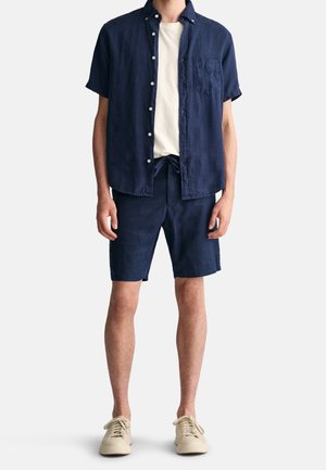 Chemise à manches courtes bleu marine avec boutonnière et poche, associée à un short assorti. T-shirt de couleur claire en dessous, complété par des baskets beiges.