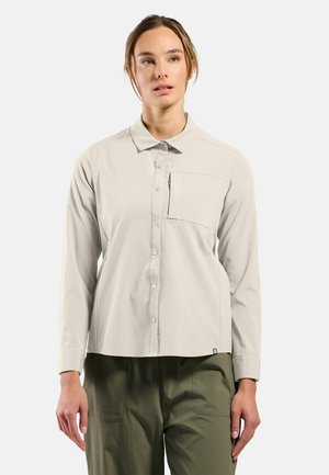 Femme portant une chemise gris clair à boutons avec une poche poitrine et un pantalon vert olive, debout devant un fond uni.
