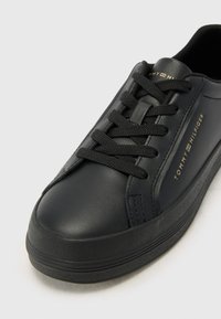 Baskets en cuir noir avec lacets plats noirs, bout rond, surface lisse, semelle en caoutchouc et logo "Tommy Hilfiger" doré sur le côté.