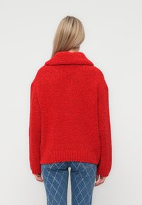 Rood, dikgebreid sweater met een brede kraag en verlaagde schouders, met een losvallende pasvorm en geribde zoom. Gecombineerd met blauwe jeans met een diamanten patroon.