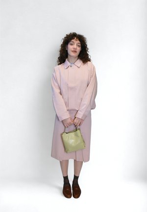 Svetloružová bunda s golierom, v kombinácii so zodpovedajúcou sukňou. Drží malú zelenú kabelku. Hnedé topánky, tmavé ponožky. Jednoduché pozadie.