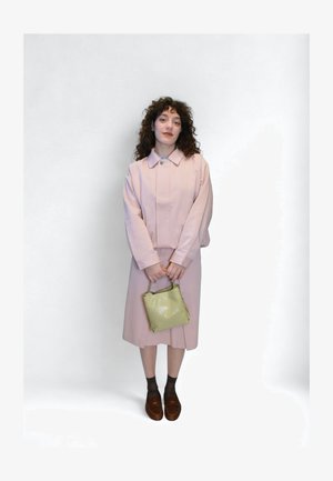 Veste rose clair avec col, associée à une jupe assortie. Tenant un petit sac à main vert. Chaussures marron, chaussettes foncées. Fond uni.