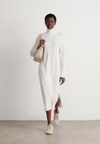 Robe tricotée blanche avec col montant et fentes sur les côtés ; modèle associé à un sac beige et des baskets beiges ; texture lisse et silhouette décontractée.