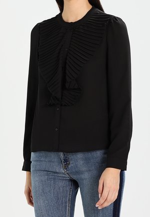 Femme portant un chemisier noir à manches longues avec un large volant plissé sur le devant, associé à un jean bleu.