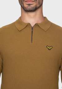 Gestikte polo sweater in mosterdgeel met een textuurstof. Heeft een kwart-rits kraag en een geborduurd geel logo op de borst.