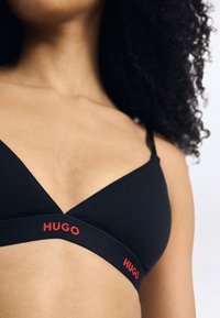 Svart trådløs bh med trekantet form, laget av mykt stoff og med en kontrastfarge rød "HUGO"-logo på båndet. Justerbare stropper inkludert.
