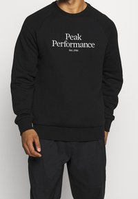 Sudadera de cuello redondo negra con puños y dobladillo acanalados. Presenta el texto blanco "Peak Performance Est. 1986" en el frente. Textura de tela suave.