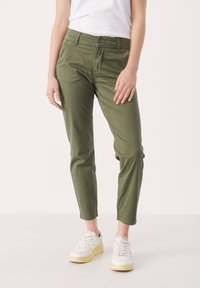 Chinos vert olive en tissu léger, avec une coupe slim, une texture subtile et une fermeture à boutons. Portés avec des baskets blanches.