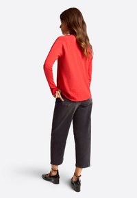 Rotes Langarmshirt aus weichem Stoff mit runder Säume. Kombiniert mit schwarzen, knöchellangen Hosen und schwarzen Blockabsatzschuhen, von hinten betrachtet.