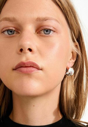 Sudraba stīpas auss auskars ar teksturētu dizainu, kurā ir izliektas formas. Modelim ir taisni, gaiši brūni mati un neitrāla ādas krāsa.