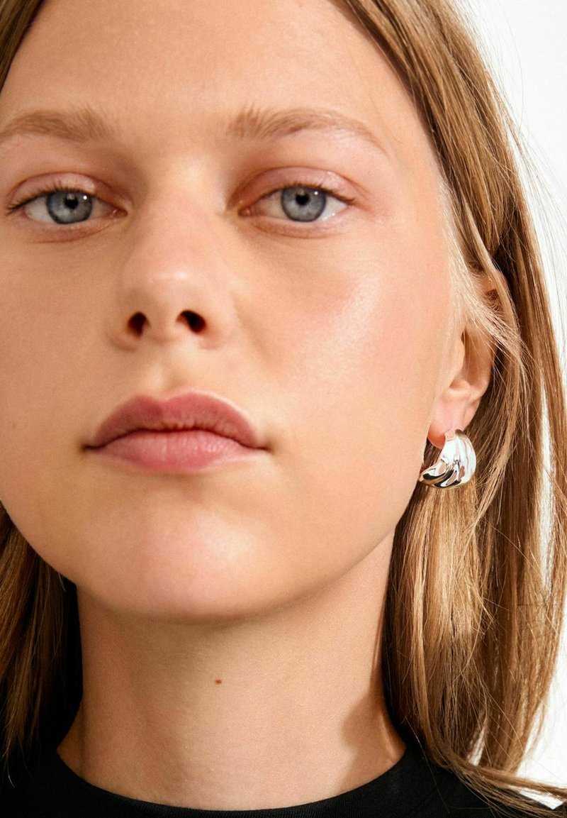 Sidabrinė lankelio formos auskaras su tekstūruotu dizainu, kuriame yra išlenktos formos. Modelis turi tiesius, šviesiai rudu plaukus ir neutralią odos spalvą.