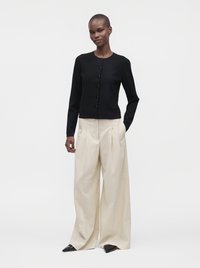 Woman wearing a black buttoned cardigan and wide beige trousers with pointed black shoes, standing against a plain light background.