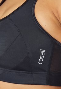 Svart sports-bh laget av glatt stoff, med mesh-paneler, sømdetaljer og en hvit logo på venstre side.
