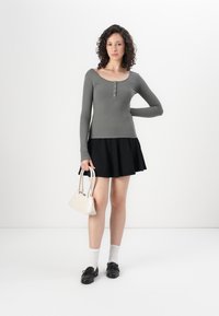 Siva rebrasta bluza s dolgim rukavima in henley gumbi, v kombinaciji z črno široko krilo. Model drži majhno bež torbico, oblečen ima bele nogavice in črne čevlje.