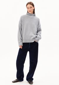 Femme portant un pull col roulé gris, un pantalon large bleu foncé et des chaussures noires à clous, se tenant avec une main dans sa poche.
