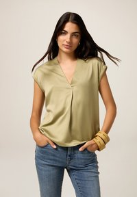 Donna con lunghi capelli scuri indossa una blusa satinata verde chiaro a maniche corte, jeans blu e grandi bracciali gialli, in piedi con le mani nelle tasche.