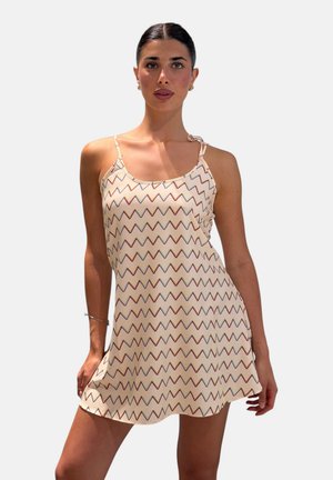 Donna con capelli scuri indossa un abito crema senza maniche con motivo a zigzag multicolore, in piedi con una mano che tiene l'orlo dell'abito.