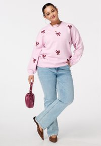 Jonge vrouw in een roze trui met rode strikken, lichtblauwe spijkerbroek, luipaardprint hakken, met een kleine bordeauxrode handtas.