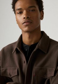 Brązowa kurtka o jodełkowym splocie z zapinanym na guzik kołnierzem, dwoma przednimi kieszeniami oraz czerwonym akcentem w postaci logo. Pod spodem widoczna jest czarna, teksturowana koszula.