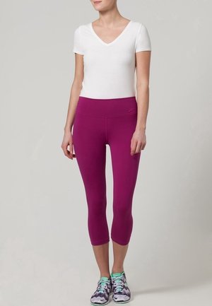 Mujer con una camiseta blanca de manga corta y cuello en V, leggings morados cortos y zapatillas deportivas moradas y blancas, de pie frente a un fondo liso.