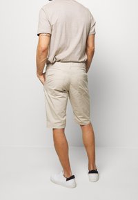 Mężczyzna w beżowych szortach, jasnym beżowym t-shircie i białych trampkach z czarnymi akcentami, stoi z rękami w kieszeniach, plecami zwrócony w stronę widza.