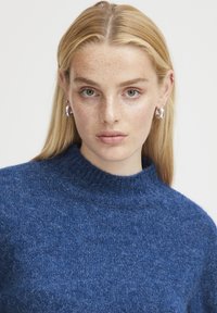 Tmavomodrý pletený sveter s rebrovaným vysokým golierom. Mäkká textúra a útulný vzhľad, nosený so striebornými kruhovými náušnicami.