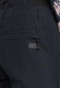 Zwarte stoffen broek met een gladde textuur, voorzien van een zijzak met een rits en een label met de tekst "ROX" op de achterkant.